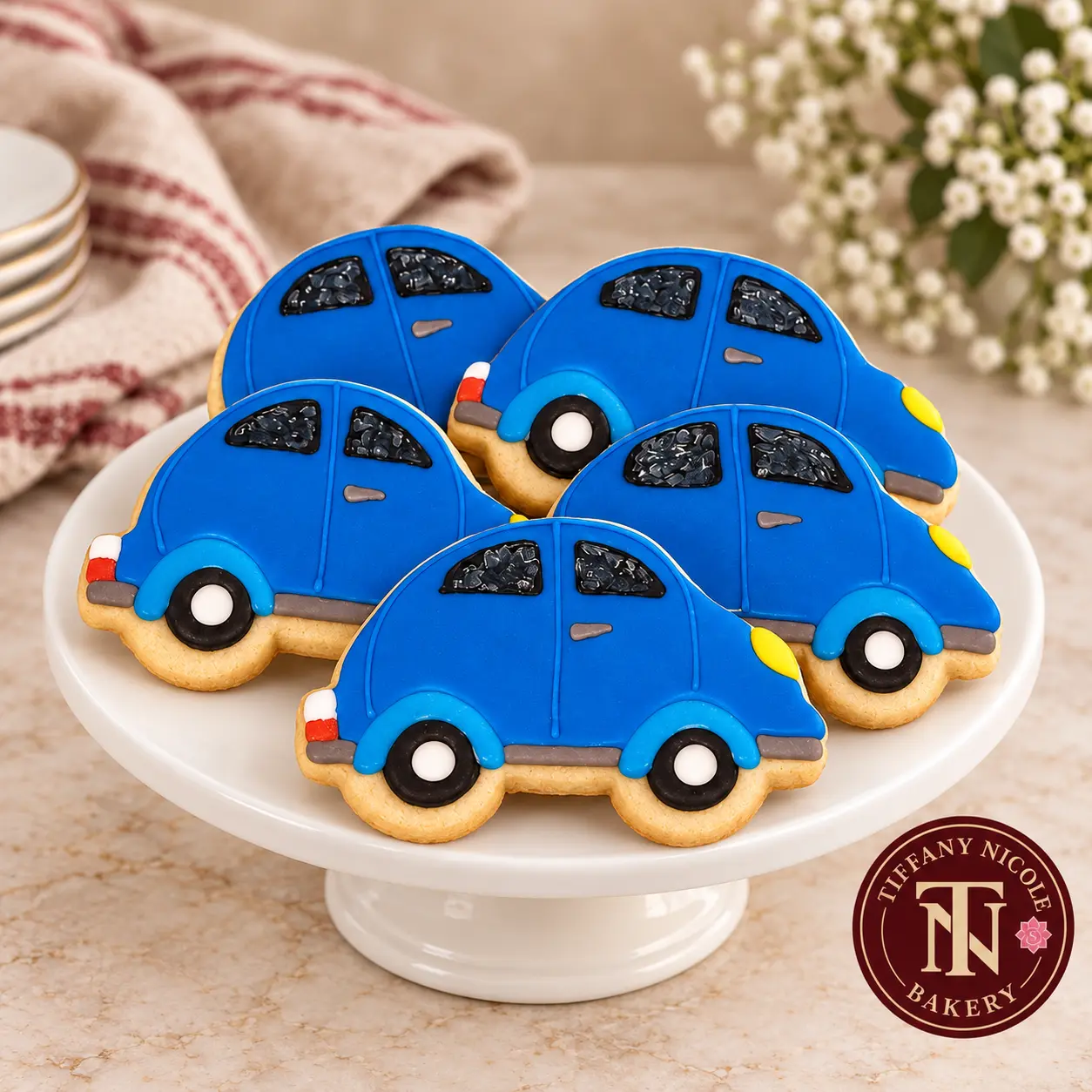 Blue car-shaped decorated sugar cookies on a white cake stand with the Tiffany Nicole Bakery logo