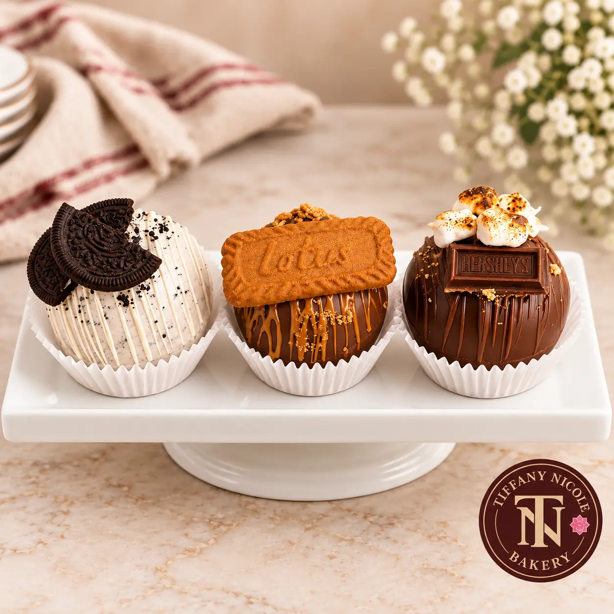 Oreo, Biscoff and s'mores hot cocoa bombs on a white dessert stand with the Tiffany Nicole Bakery logo